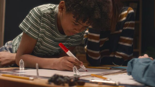 Medium shot of African American boy leaning on table and drawing location map for table game while his diverse friends watching him at home in evening