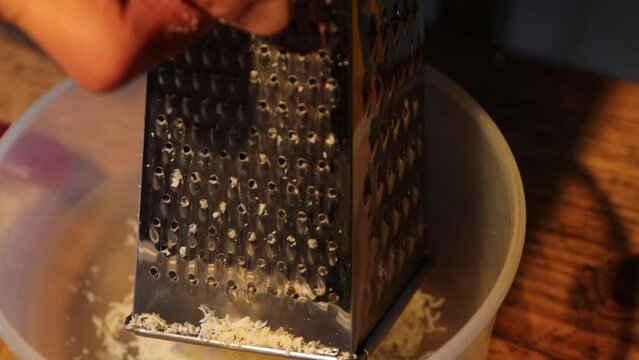 Man is rubbing parmesan into a plastic bawl on a wooden table. Hands rub cheese on a metal grater. Cooking a meal with cheese. Working in the kitchen in the evening. Dark background.