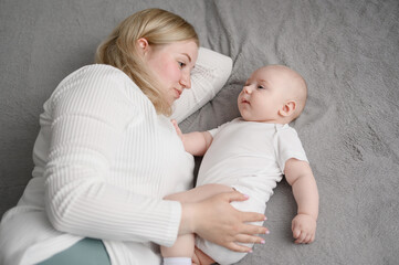 Mom woman lies on bed, holding her baby close to her, showing her love so that child feels comfort and care. Motherhood, infancy.