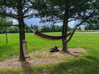 A hammock on a tree at the park with a longboard and a backpack beside it