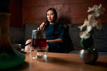 Self-confident Asian woman sitting on sofa smoking fresh and strong hookah