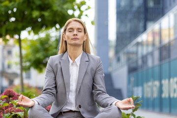 Tired young business woman sitting on bench outside office work building in lotus position with...
