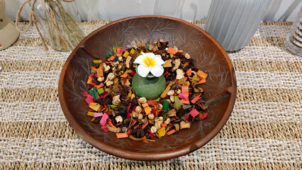 Decorative centerpiece with a white frangipani flower on a green ceramic base surrounded by colorful dried potpourri in a brown leaf etched bowl on a woven mat, creating a cozy and inviting atmosphere