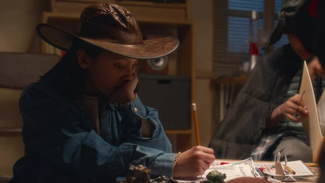 Side shot of bored Hispanic girl wearing cowgirl costume filling in character sheet during role playing game at home in evening