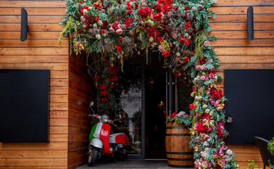 An enchanting floral archway bursts with a cascade of red and pink blooms, framing the entrance to a rustic wooden venue, where a classic scooter stands as a charming accent