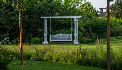 Fototapeta premium A tranquil white garden swing bed nestled among lush greenery in a peaceful garden, offering a picturesque spot for relaxation amid blooming roses and verdant shrubs