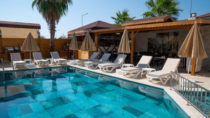 a serene poolside area with crystal-clear blue water. Surrounding the pool are white sun loungers, each paired with a folded beige umbrella. A wooden pergola with a bar can be seen in the background