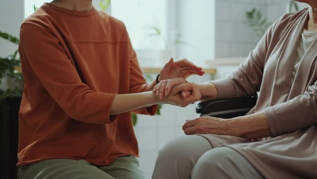 Woman holding hand of senior grandmother sitting in wheelchair and having talk with her while visiting at home. Midsection shot