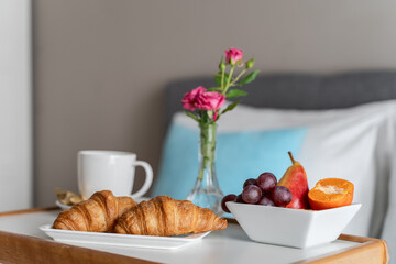 a wooden tray with food on the bed. Fruits, croissants, a white mug with coffee or tea and pink roses on a tray. A bed with white and blue pillows. Breakfast in bed