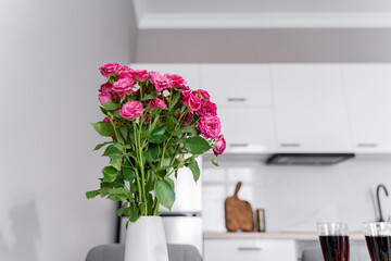 A white vase with small pink roses in the kitchen. White modern kitchen.