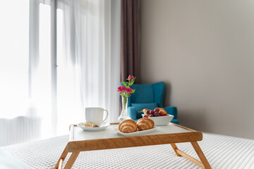 There are fruits on a wooden tray in a white plate, croissants on a plate, roses in a vase and a mug of coffee. The tray is on the bed. Breakfast in bed. The blue armchair in the bedroom.