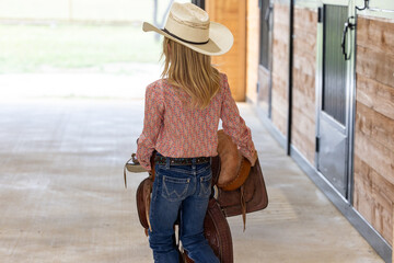 Little cowgirl carrying saddle wearing a straw cowboy hat