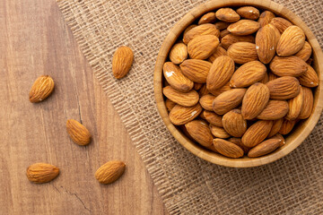 Overhead view of a bowl full of peeled almonds on a wooden surface. A piece of burlap and a few almonds lying around