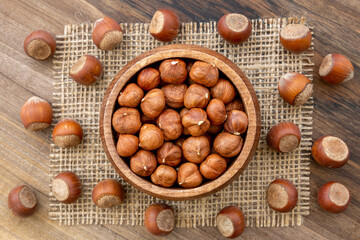 Overhead view of a bowl full of hazelnut kernels on a wooden surface. A piece of burlap and a few shelled hazelnuts lying around