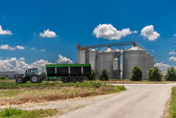 Tractor with grain harvesting trailer transits in front of steel grain storage silos on blue sky in the Po Valley in the province of Cuneo, Piedmont, Italy