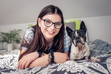 young girl with glasses and a Boston Terrier dog lying on a bed, both smiling at the camera. The cozy bedroom features a patterned blanket and a plant, ideal for themes of companionship and relaxation