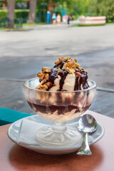 Vanilla ice cream with chocolate topping and walnuts in a glass on the table, outdoor cafe area on a sunny summer day