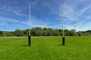 A pair of rugby goal posts stand prominently at the center of a lush, green field under a blue sky. In the background, trees line the edge of the playing area in, Cottingley, UK