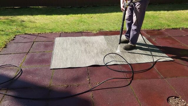 Washing carpet on rubber flooring in backyard. Man cleaning stone garden paths with high pressure power washer. Taken from hand. High quality 4k footage
