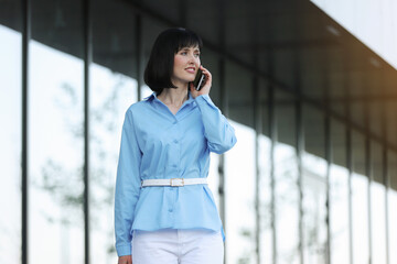 Successful elegant businesswoman entrepreneur or CEO in blue shirt and white pants smiles and talks on smartphone with partners or clients while walks by the office building outdoor. Business concept