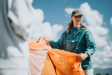 A young woman setting up an orange tent outdoors on a sunny day. She is dressed casually in a blue...