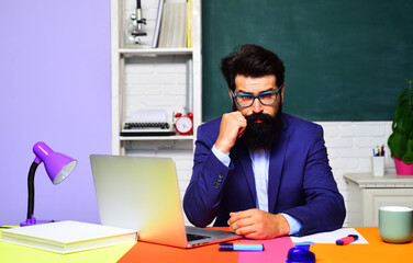 Serious male university teacher working with laptop computer in classroom. E-learning and education. Online test or exam in college. Bearded student sitting at desk with notebook at school lesson.