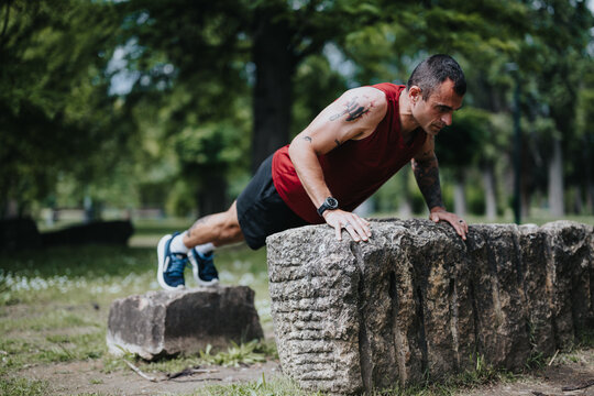 Focused athletic man with tattoos performing push-ups on a rustic stone bench in a lush green park, exemplifying dedication and strength in his fitness routine.