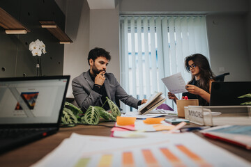 In a well-lit office, a male and female colleague discuss documents earnestly, surrounded by technology and business charts.
