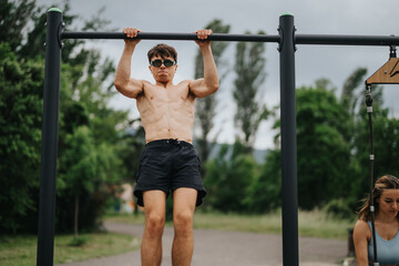 Fototapeta premium Man exercising by doing pull-ups on a bar in an outdoor park setting, focusing on strength training and fitness.