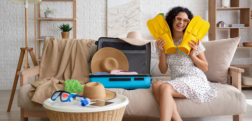 Young African-American woman unpacking suitcase with travelling accessories and flippers on sofa in hotel room © Pixel-Shot