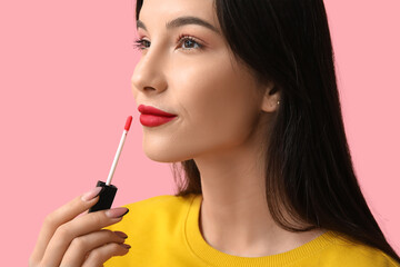 Fototapeta premium Beautiful young woman applying red lipstick on pink background