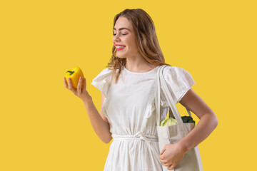 Pretty young woman with fresh vegetables in shopper bag on yellow background