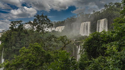 Beautiful cascades of tropical waterfalls. White foaming streams fall from the ledges of the rocks. Splashes, fog all around. Lush green vegetation. Clouds in the blue sky. Iguazu Falls. Argentina.