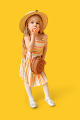 Cute little girl in hat with stylish wicker bag blowing kiss on yellow background