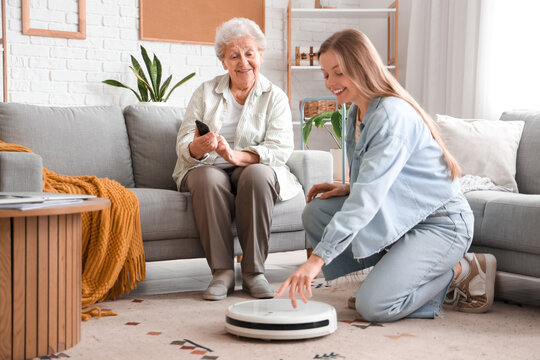 Young woman showing her grandmother how to use robot vacuum cleaner at home