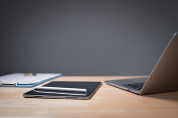 A laptop and tablet sit on a desk with a clipboard and pen. The desk is cluttered with papers and the laptop is on a wooden surface
