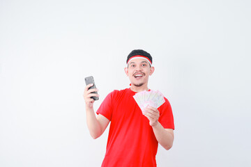 Cheerful young Asian man in casual t-shirt holding mobile phone and money banknotes isolated on grey background. indonesian independence day celebration concept