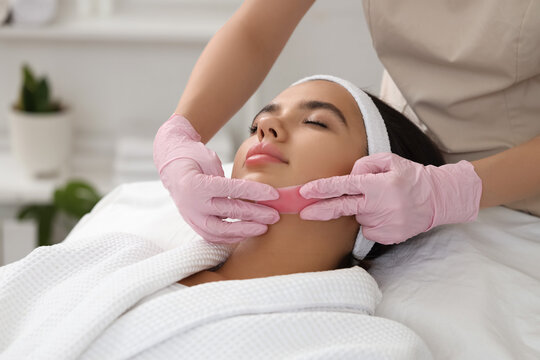 Beautician with wax strip removing woman's hair on face in salon, closeup