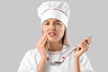 Young female chef with ladle suffering from tooth ache on grey background
