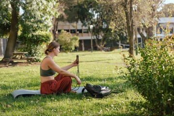 Fototapeta premium Middle aged woman, phone and network outdoor for communication, social media and reading news app in summer park. Lady taking selfie while sitting on yoga mat. Unity with nature, sport yoga concept.