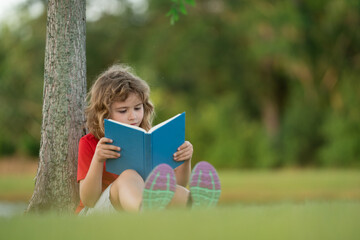 Outdoor portrait of a cute little kid reading a book in summer park. Blond kid boy sitting on grass and reading book. Summer weekend in park. Schoolboy child study with book, learning. Book store.