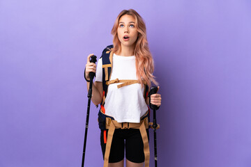 Teenager girl with backpack and trekking poles over isolated purple background looking up and with surprised expression