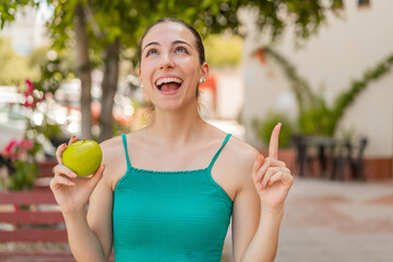 Young pretty woman with an apple at outdoors intending to realizes the solution while lifting a finger up