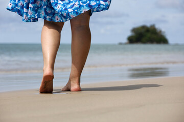 Barefoot woman in summer dress walking by the sand on sea waves and island background. Female legs on a coast, beach vacation