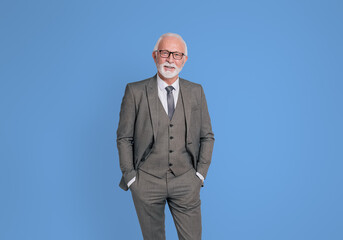 Portrait of confident senior businessman with hands in pockets standing on isolated blue background