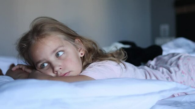 A little blonde girl lies in her cozy bed on her stomach under a warm and cozy blanket and looks towards the concept of rest, relaxation and awakening or daytime sleep in children.