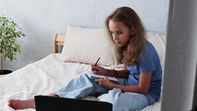 A charming teenage girl takes notes in a notebook during online lectures, which she watches on her laptop while in a cozy home environment