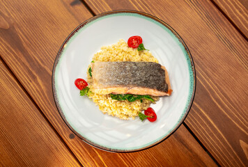 Grilled baked salmon garnished with bulgur, tomatoes, herbs and lemon. Fish dish, grilled salmon steak on a wooden table.