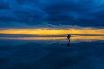 Fototapeta premium A lone traveler with a backpack walks across calm, reflective water under dramatic skies at sunset. Adventurous, peaceful, and inspiring.