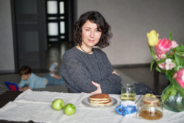 Positive middle age women sitting the the table at home with tea and pancakes. Beautiful lade looking at the camera, smiling, wearing casual clothing. High quality photo